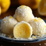 Homemade lemon truffles coated in powdered sugar with lemon zest, one bitten to show a creamy lemon center, on a white plate.