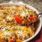 Homemade stuffed pepper casserole topped with melted cheddar and mozzarella, ground beef, rice, red and green bell peppers, and fresh herbs, with a wooden spoon lifting a cheesy serving.
