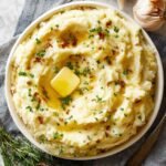 Homemade mashed Potatoes Recipe topped with melting butter, sprinkled with parsley and crispy browned bits, served on a gray napkin with garlic and rosemary nearby.