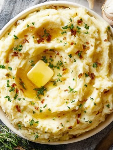 Homemade mashed Potatoes Recipe topped with melting butter, sprinkled with parsley and crispy browned bits, served on a gray napkin with garlic and rosemary nearby.