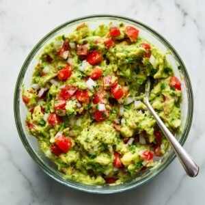 Chunky homemade guacamole with avocado, diced tomatoes, onion, cilantro, and black pepper in a glass bowl with a spoon, on a white marble countertop in natural light.