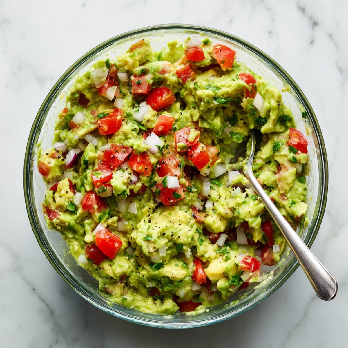 Chunky homemade guacamole with avocado, diced tomatoes, onion, cilantro, and black pepper in a glass bowl with a spoon, on a white marble countertop in natural light.