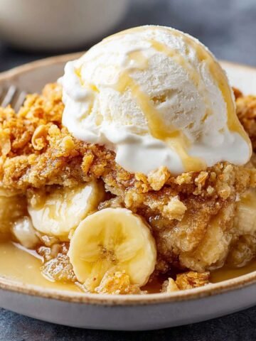 Banana Cobbler Recipe topped with vanilla ice cream and syrup, with visible banana slices and a golden crumb topping. A spoon rests in the bowl, and a whole banana is in the background.