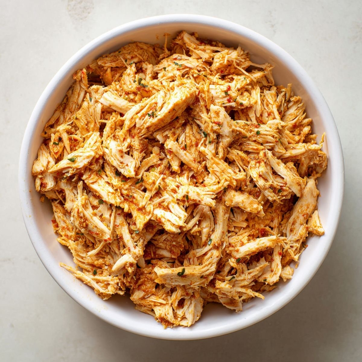 Seasoned shredded chicken filling in a white bowl, coated with spices and herbs, ready to be used for homemade chicken flautas.
