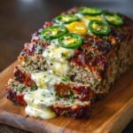 Freshly sliced Jalapeño Popper Meatloaf on a wooden board, filled with melted cheese oozing from the center, topped with golden cheddar, sliced green jalapeños, and finely chopped parsley, with a crispy, caramelized crust.
