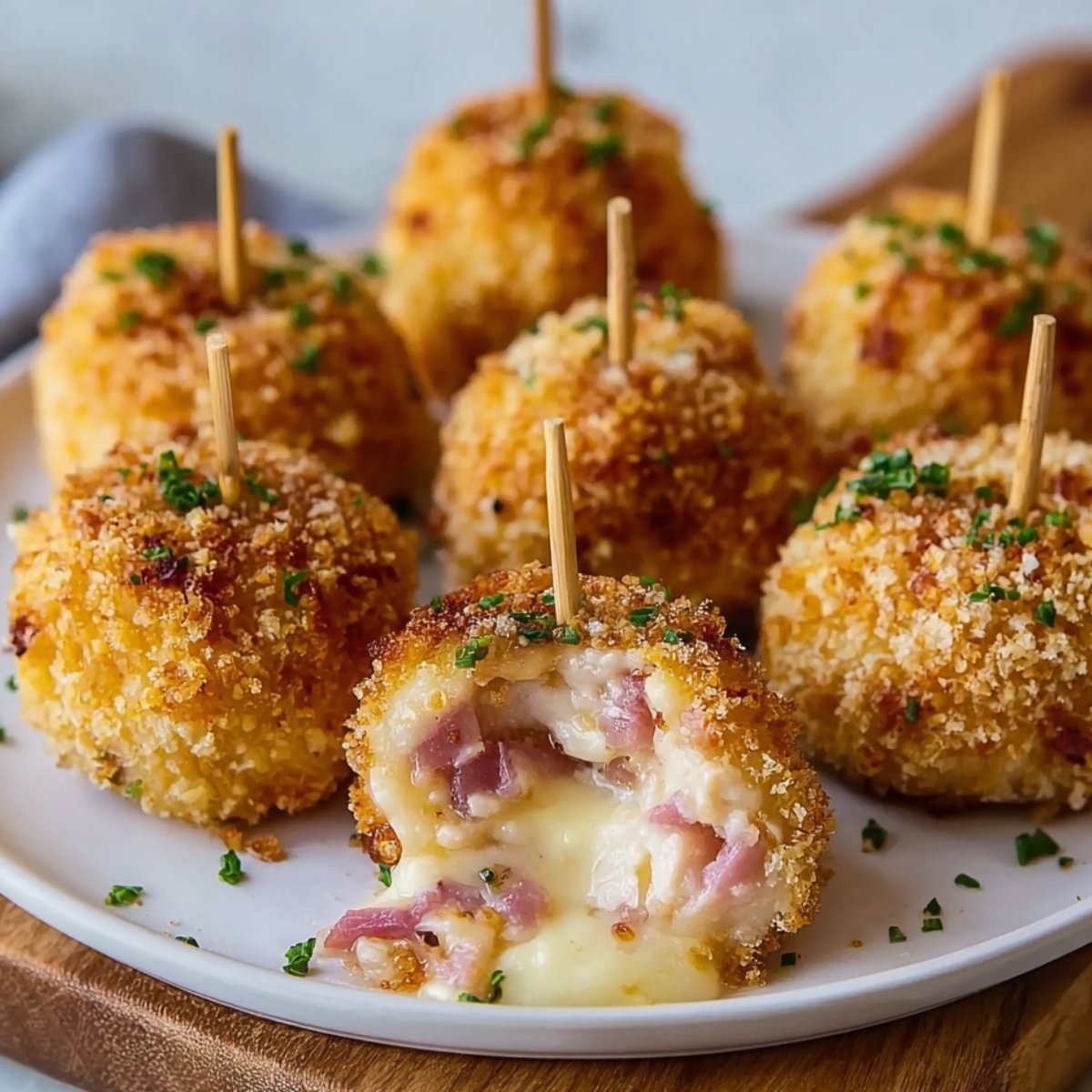 Crispy homemade Chicken Cordon Bleu Bites on a white plate with skewers, topped with parsley. One bite is cut open, showing melted cheese and ham inside.