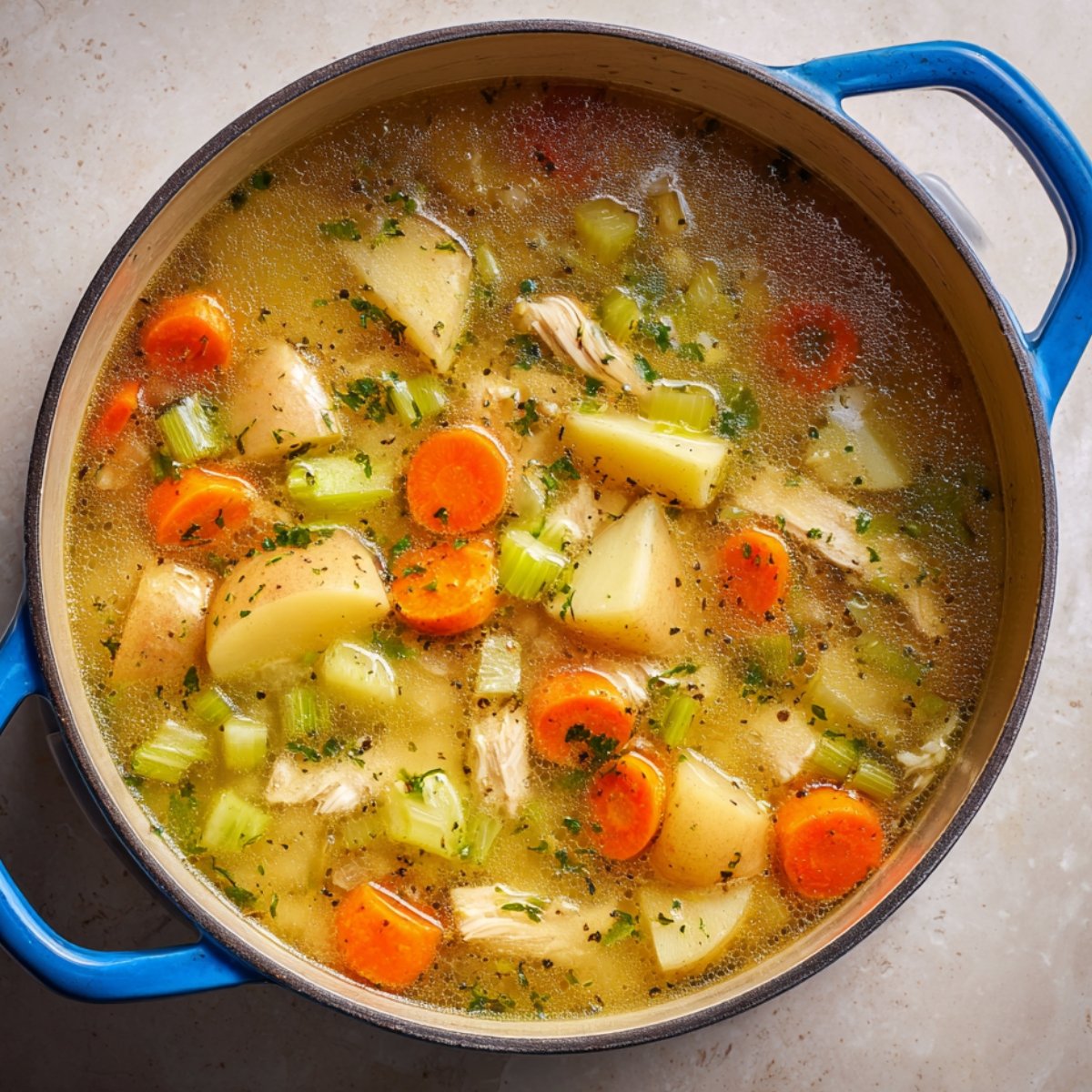 Homemade chicken soup in a blue Dutch oven with golden broth, chunks of potato, sliced carrots, celery, and shredded chicken, topped with fresh parsley — warm, hearty, and freshly made.