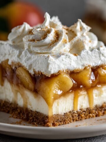 Homemade Caramel Apple Eclair Cake with a graham cracker crust, creamy filling, caramelized apples, and whipped cream topped with cinnamon, served on a beige plate with a blurred red apple in the background.
