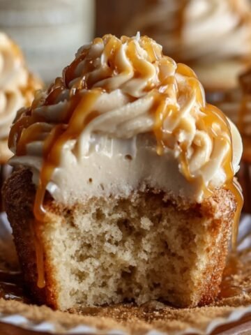 Homemade Harry Potter butterbeer cupcakes with creamy caramel-drizzled frosting, one bitten to show soft, fluffy inside, resting on a book with more cupcakes blurred in the background under warm, cozy lighting.