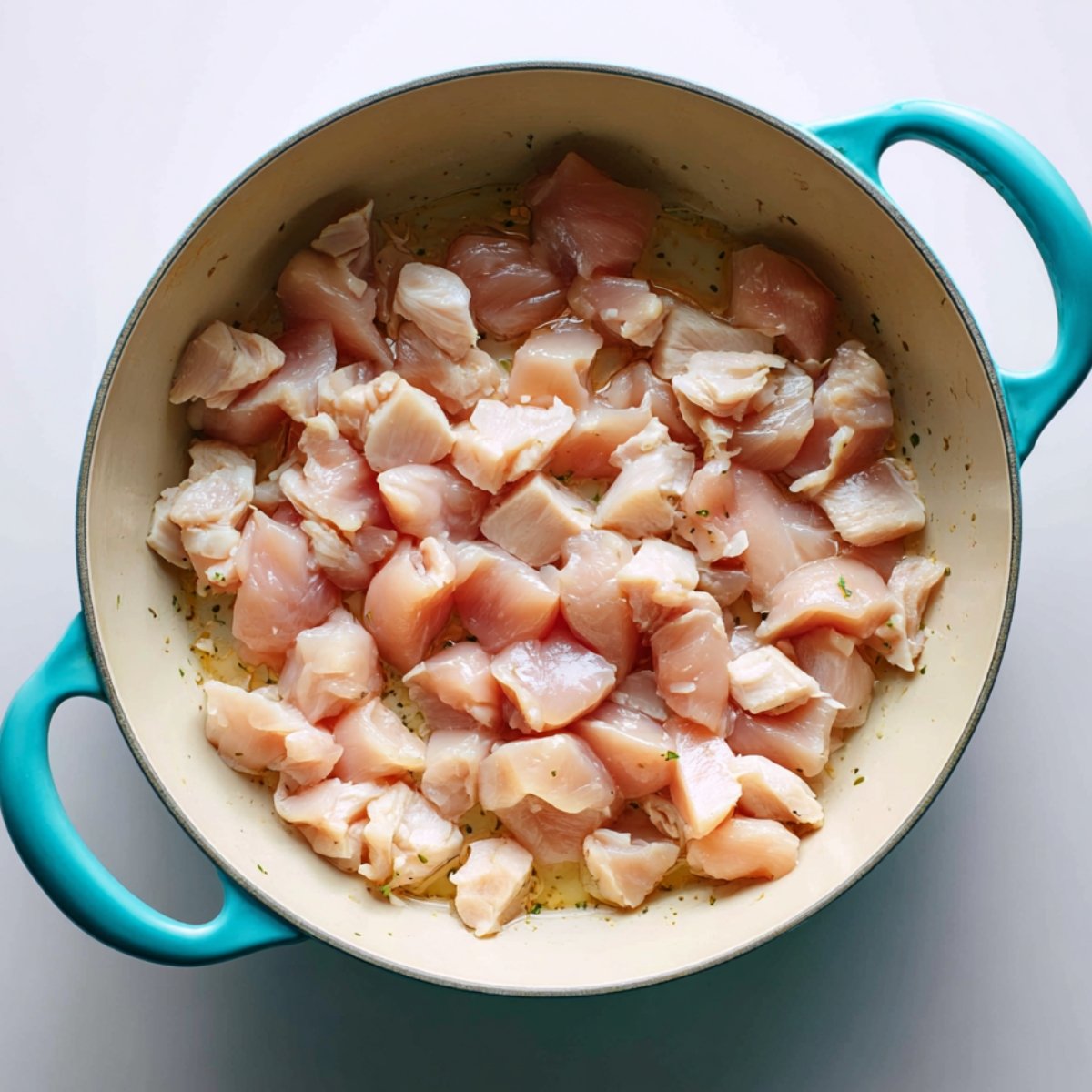 Raw diced chicken breast pieces spread in a large cream-colored Dutch oven with teal handles, ready to be cooked for homemade Bisquick chicken and dumplings.