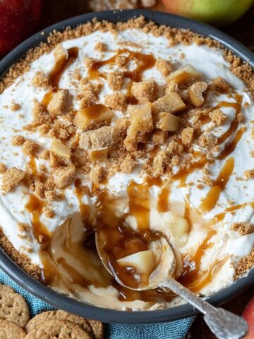 Homemade caramel apple cheesecake dip topped with caramel drizzle, crumbly brown sugar streusel, and diced apples in a round dish, surrounded by apples and graham crackers.