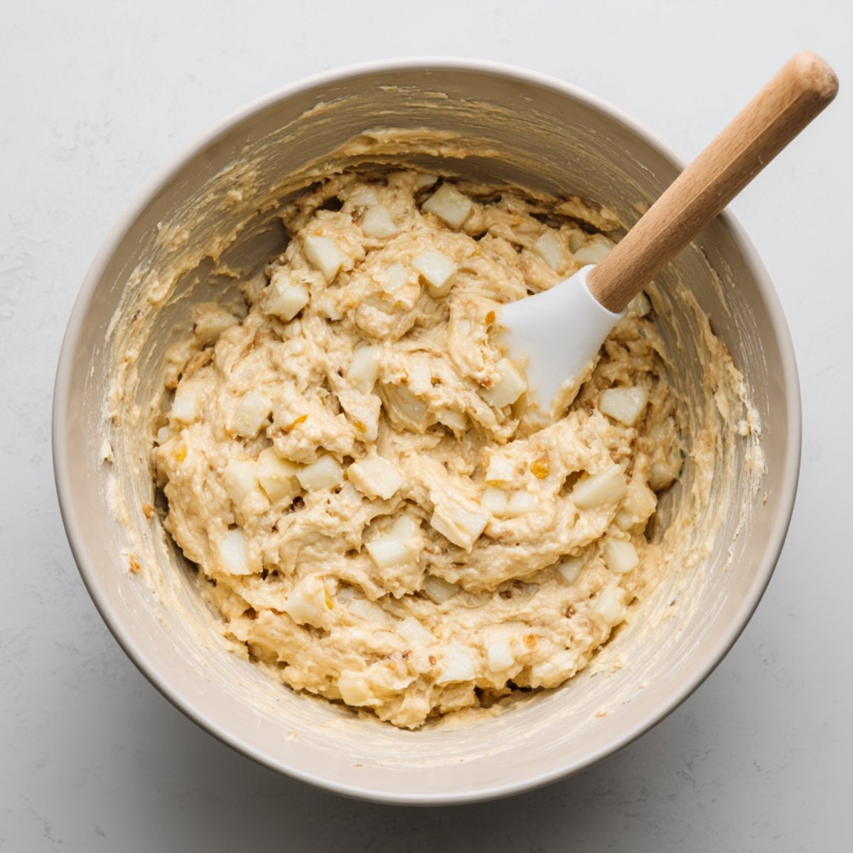 Mixing bowl filled with thick apple fritter batter speckled with diced apples, stirred with a white spatula with a wooden handle on a light kitchen countertop.