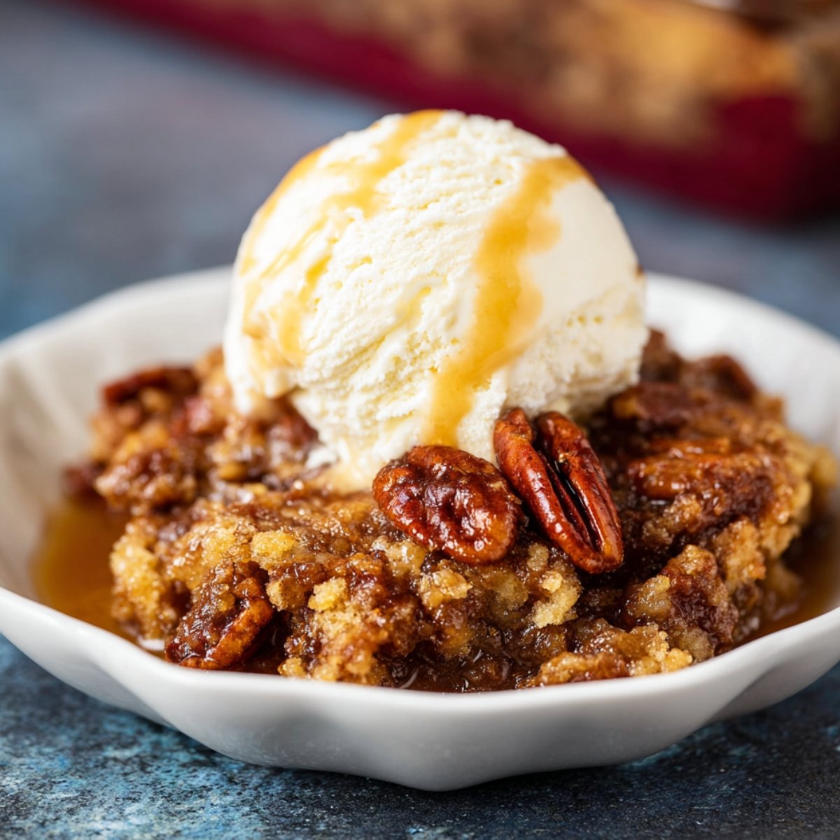 Pecan Pie Cobbler topped with a scoop of melting vanilla ice cream and caramel drizzle in a white scalloped bowl.