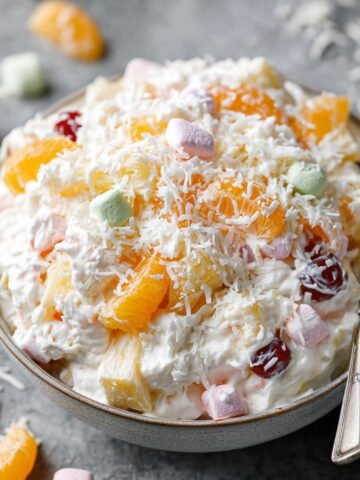 Homemade ambrosia fruit salad in a bowl, topped with shredded coconut, mandarin orange slices, cherries, and pastel mini marshmallows.