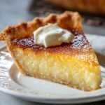 Slice of homemade buttermilk pie with a golden, flaky crust, creamy filling, and topped with a dollop of whipped cream, served on a white plate.