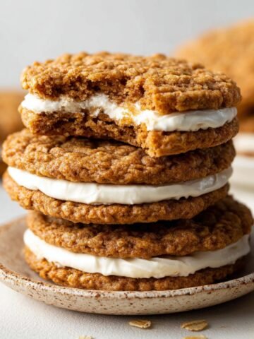 Homemade Oatmeal Cream Pies with one showing a bite taken out, revealing the creamy filling between the oatmeal cookies. The cookies are golden brown and placed on a simple ceramic plate.