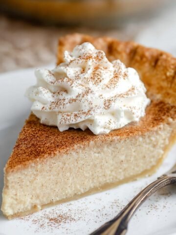 Slice of Vanilla Cinnamon Pie with whipped cream and a dusting of cinnamon, served on a white plate with a vintage fork.