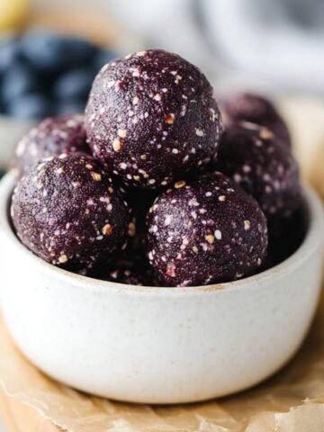 Blueberry muffin energy balls stacked in a small white bowl, showing a dark purple no-bake snack texture.