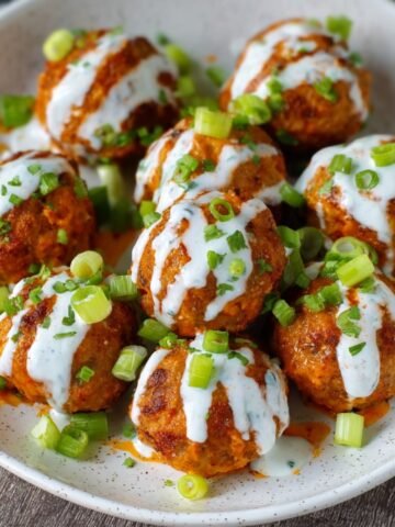 A close-up shot of cooked Buffalo Chicken Meatballs covered in spicy buffalo sauce, garnished with chopped green onions and served in a bowl.