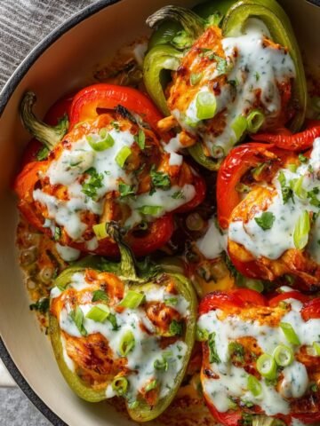 Close-up view of roasted red and green Buffalo Chicken Stuffed Peppers Recipe, drizzled with creamy sauce and garnished with sliced green onions and herbs, showing melted, slightly charred edges.