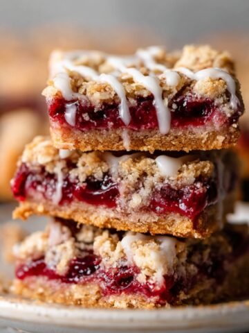 A stack of freshly baked Cherry Pie Bars with a golden, crumbly oat topping, filled with rich cherry filling and topped with a light drizzle of icing.