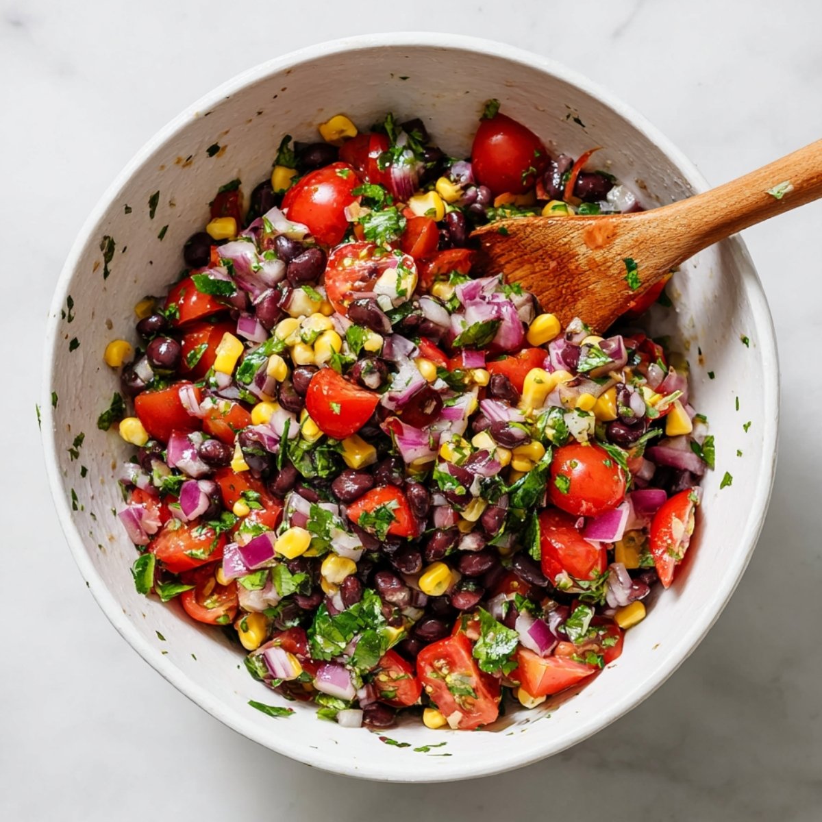 Overhead view of a bowl of black bean salsa with corn, cherry tomatoes, red onion, cilantro, and a wooden spoon.