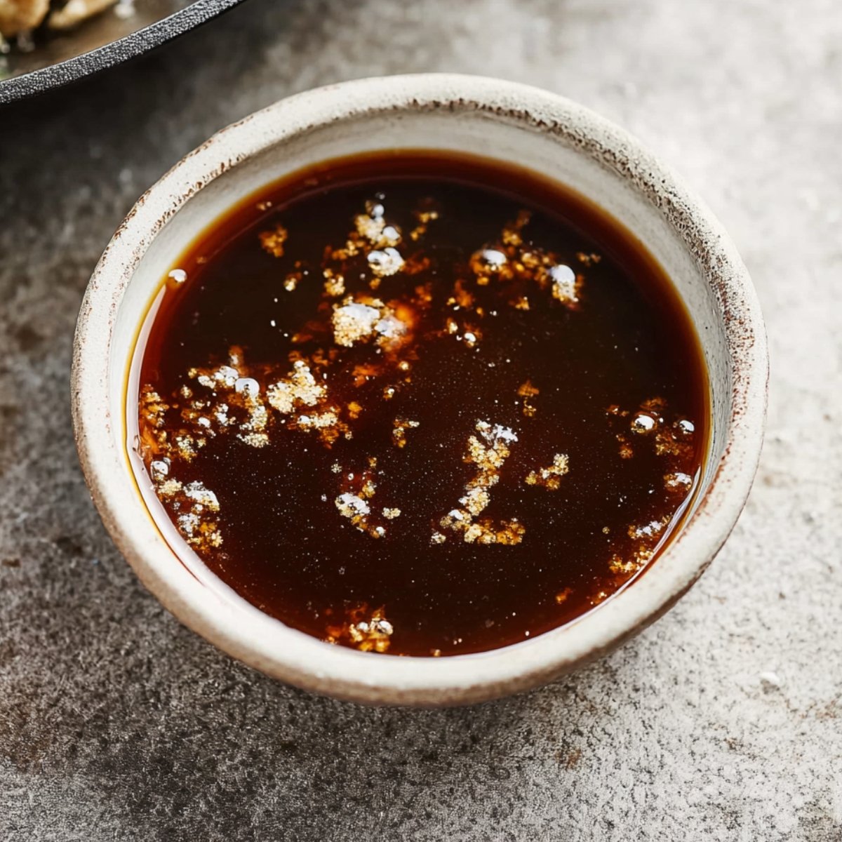 A small bowl filled with a dark, glossy sauce, likely made with soy sauce or a similar base, with small bits of seasoning visible floating on the surface.