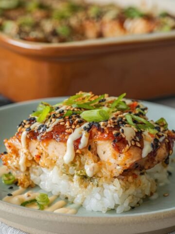 A close-up of Spicy Salmon Sushi Bake on a plate, topped with sesame seeds, green onions, and creamy sauce, with a baking dish in the background.