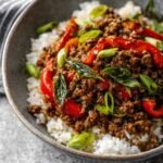 A completed dish of Thai basil beef served over white rice in a gray bowl. The dish features ground beef, red bell peppers, and fresh basil leaves, garnished with sliced green onions.