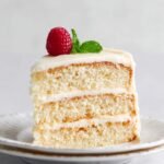 A slice of Vanilla Cake showing the layers of soft cake and frosting, topped with a raspberry and mint sprig.