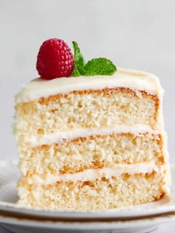 A slice of Vanilla Cake showing the layers of soft cake and frosting, topped with a raspberry and mint sprig.