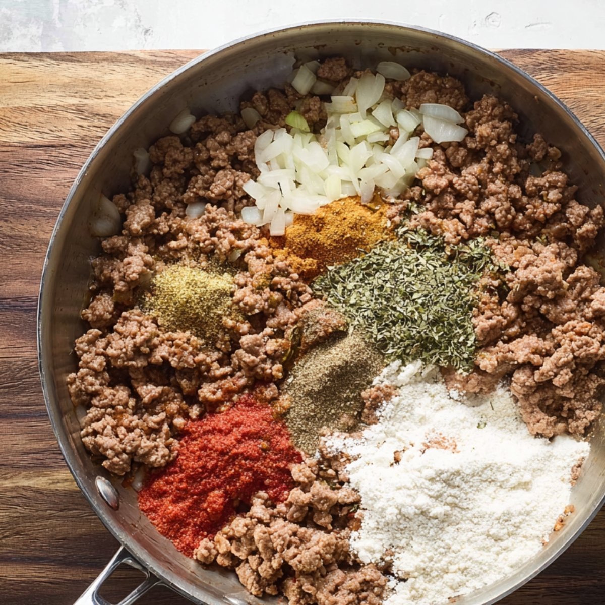 A skillet filled with ground beef, diced onions, and an array of spices like paprika, garlic powder, and oregano, along with flour to help thicken the sauce.