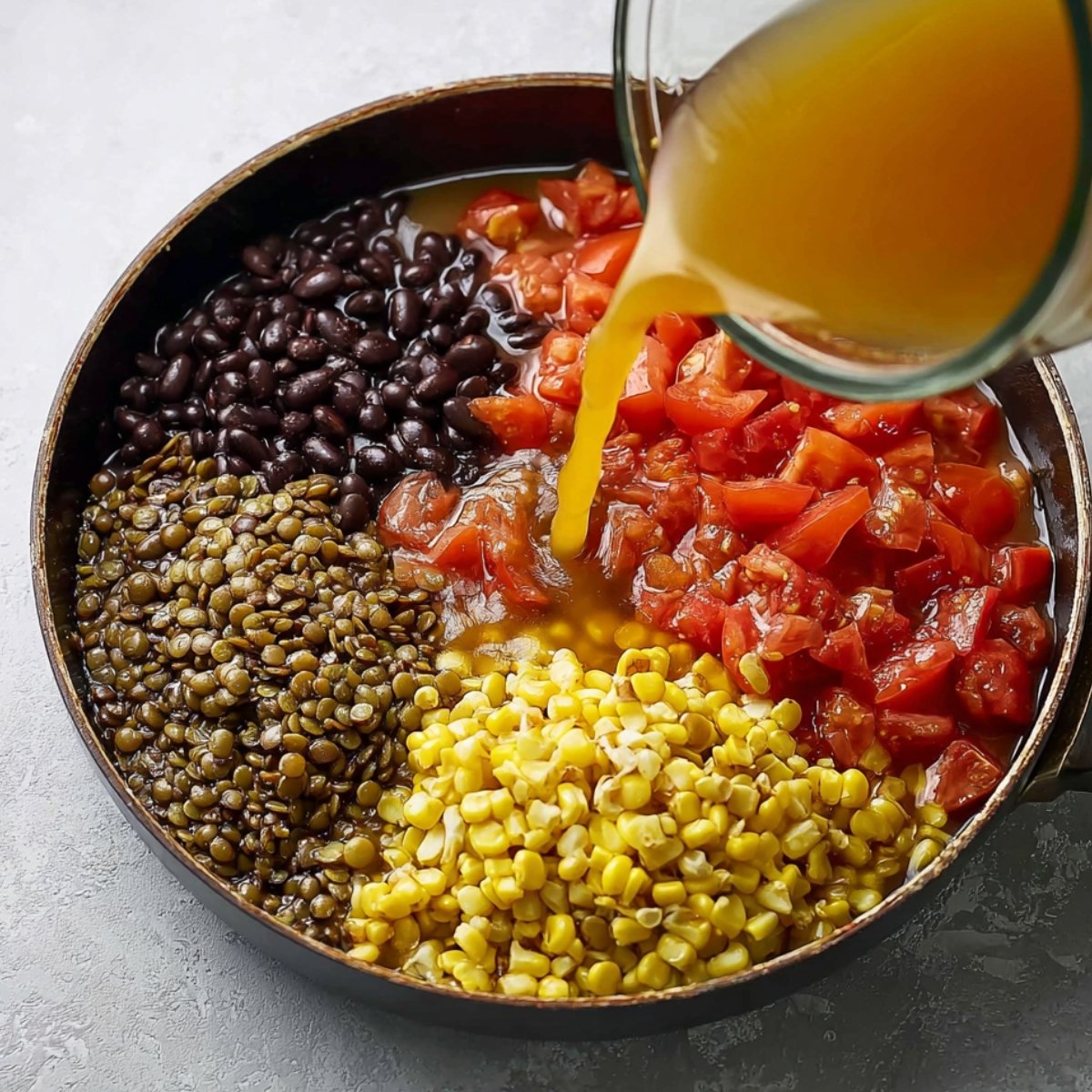 A skillet filled with black beans, corn, diced tomatoes, and lentils, with broth being poured in, preparing the base for a flavorful dish.