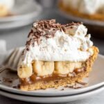 A close-up of a decadent slice of Banoffee Pie, topped with whipped cream, chocolate shavings, and a layer of banana slices on a buttery graham cracker crust.