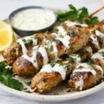 Grilled Chicken Kofta Kebabs drizzled with creamy white sauce and garnished with fresh herbs, served alongside lemon wedges and a dipping sauce.
