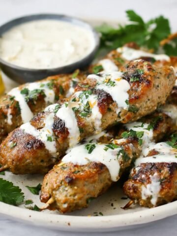 Grilled Chicken Kofta Kebabs drizzled with creamy white sauce and garnished with fresh herbs, served alongside lemon wedges and a dipping sauce.