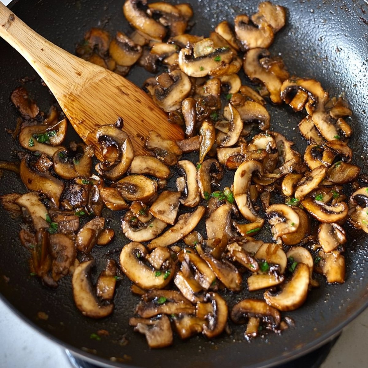 Sautéed sliced mushrooms in a pan, cooked to a golden brown with a wooden spoon in the mix.