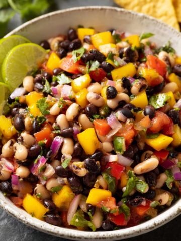 A bowl of Cowboy Caviar Recipe, featuring black beans, black-eyed peas, tomatoes, yellow bell peppers, red onions, and cilantro, garnished with lime wedges.