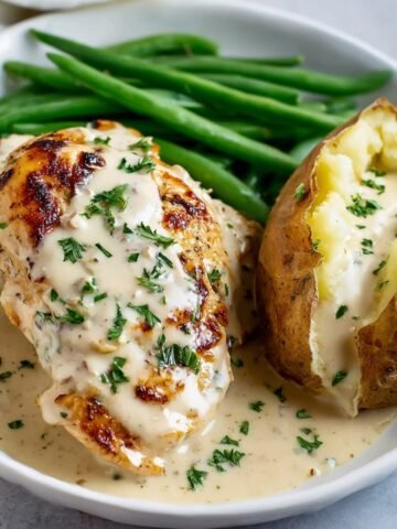 A plate Creamy Ranch Chicken covered in creamy ranch sauce, garnished with chopped parsley. The dish includes green beans and a baked potato with creamy sauce.