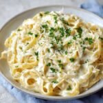 Plate of creamy Fettuccine Alfredo topped with fresh parsley and grated parmesan cheese. The pasta is rich and creamy, served on a light blue napkin.