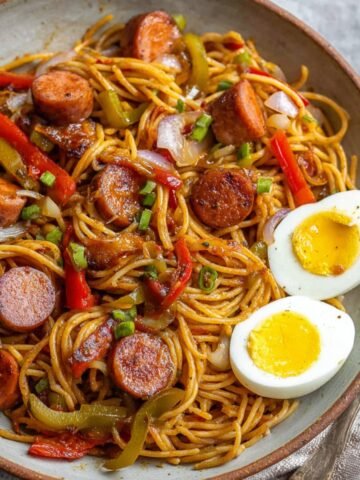 A serving of Haitian spaghetti in a rustic bowl with sliced sausages, bell peppers, onions, and soft-boiled eggs on the side. The dish is colorful and hearty, making for a comforting meal.