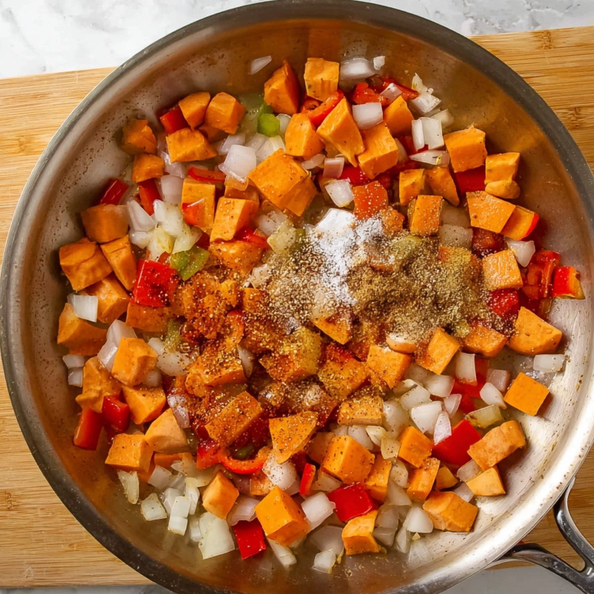 Diced sweet potatoes, onions, and bell peppers in a skillet, with seasonings like salt, pepper, and spices added on top, ready to be cooked.
