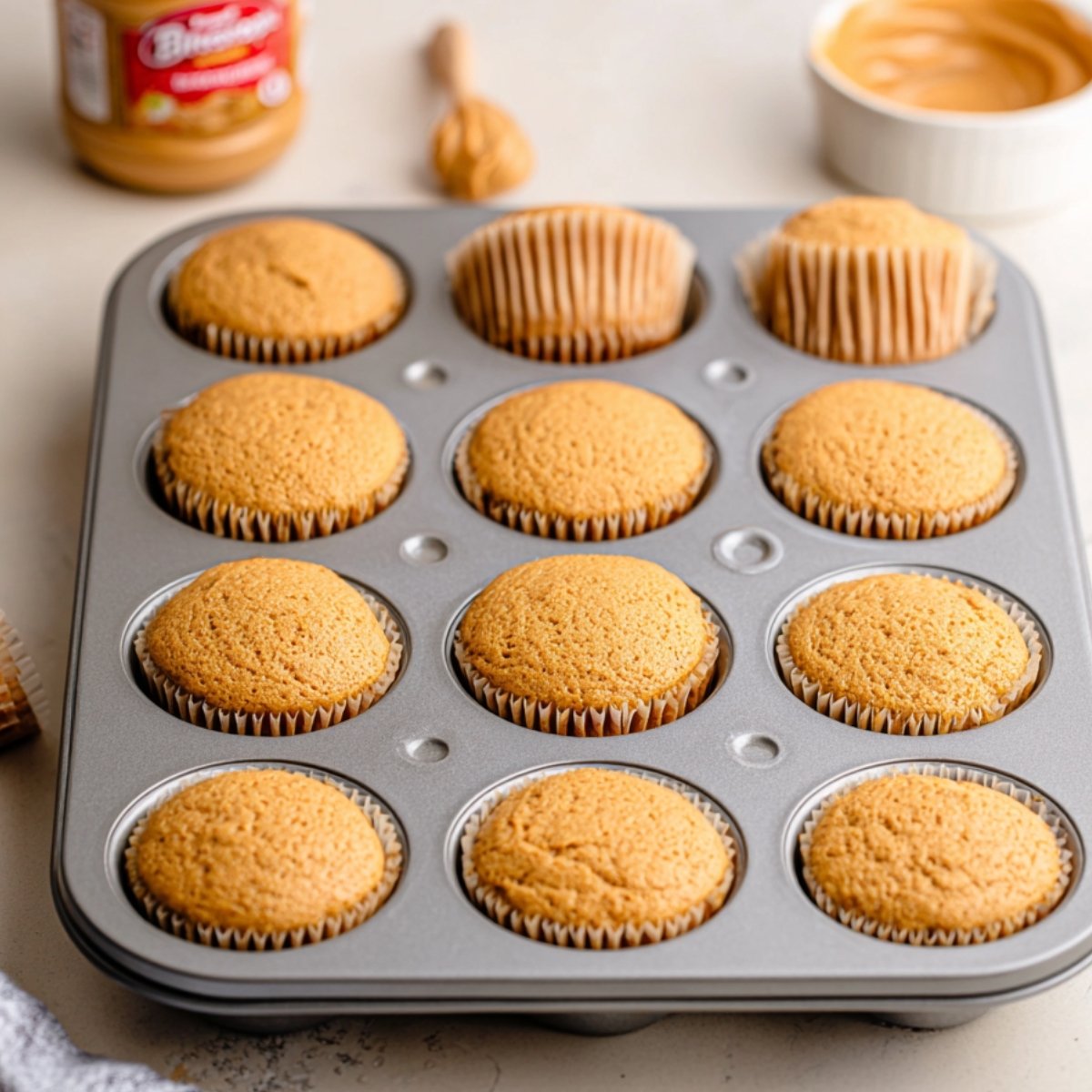 Golden Biscoff cupcakes cooling in a muffin tin, with a jar of Biscoff cookie butter nearby, waiting to be topped with frosting.