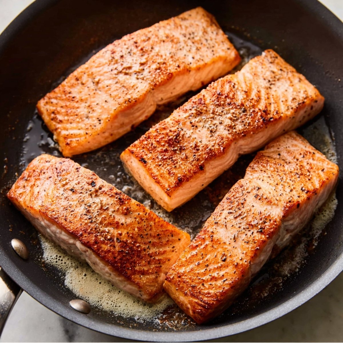 Salmon fillets searing in a skillet, lightly browned with a crispy edge, seasoned with salt and pepper.