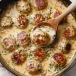 A close-up of Creamy Marry Me Chicken Meatballs in a skillet, simmering in a rich sauce, with a wooden spoon lifting one meatball, surrounded by a smooth, savory sauce.