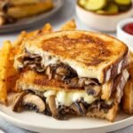 A close-up of a delicious Mushroom Patty Melt sandwich, featuring golden toasted bread, melted cheese, and sautéed mushrooms, served with crispy fries and pickles.