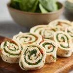 Turkey pinwheels made with tortillas, spinach, and creamy filling, neatly sliced and arranged on a wooden board, with a bowl of fresh spinach in the background.